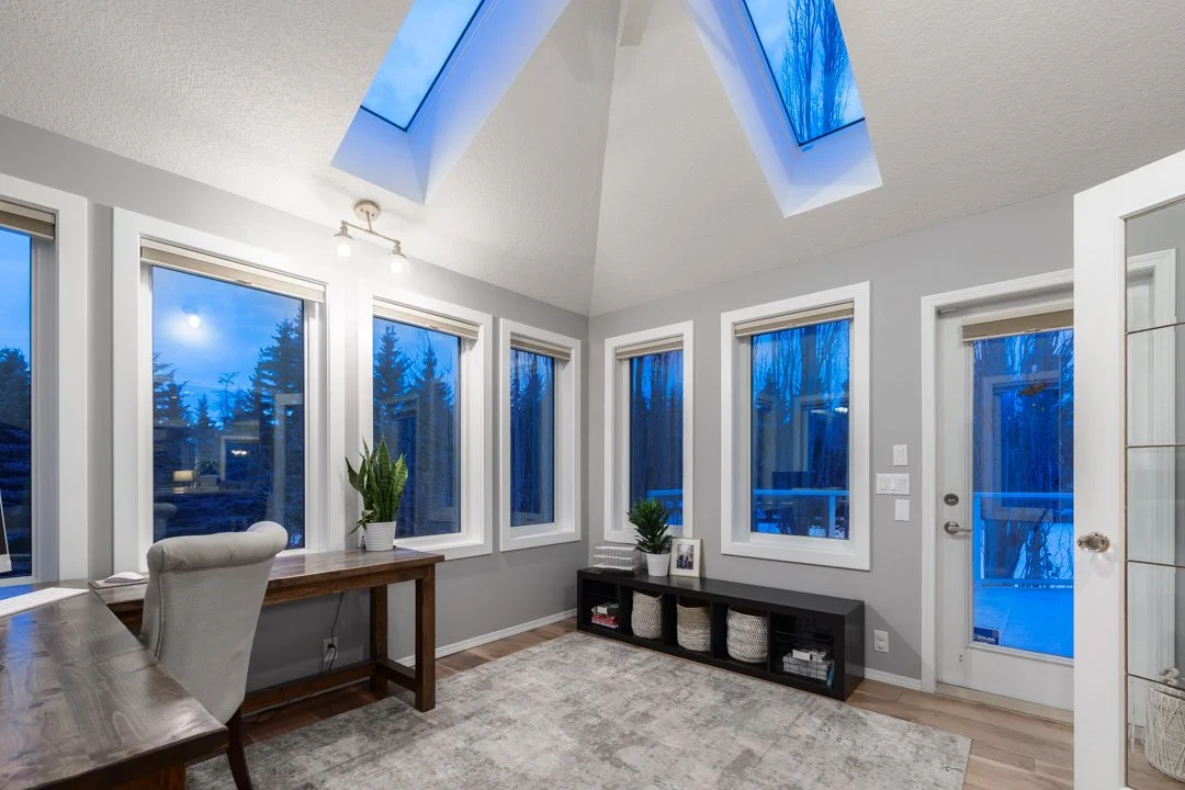 Bright sunroom with five large windows, a skylight, a gray wall, a wooden desk with a beige chair, a small black storage unit, decorative plants, and a rug on hardwood flooring.