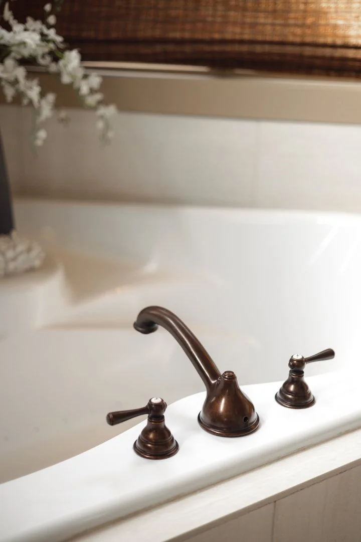 Close-up of a bronze-colored bathroom faucet and handles on a white bathtub with a wooden plank and decorative flowers in the background.