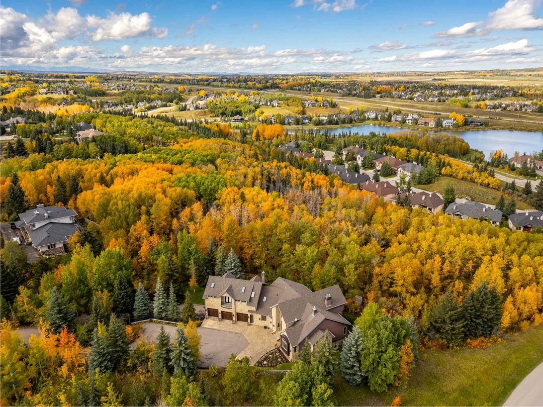 Aerial view of a suburban neighborhood surrounded by colorful fall trees, with a large house in the foreground, a lake in the mid-ground, and open fields in the background under a partly cloudy sky.