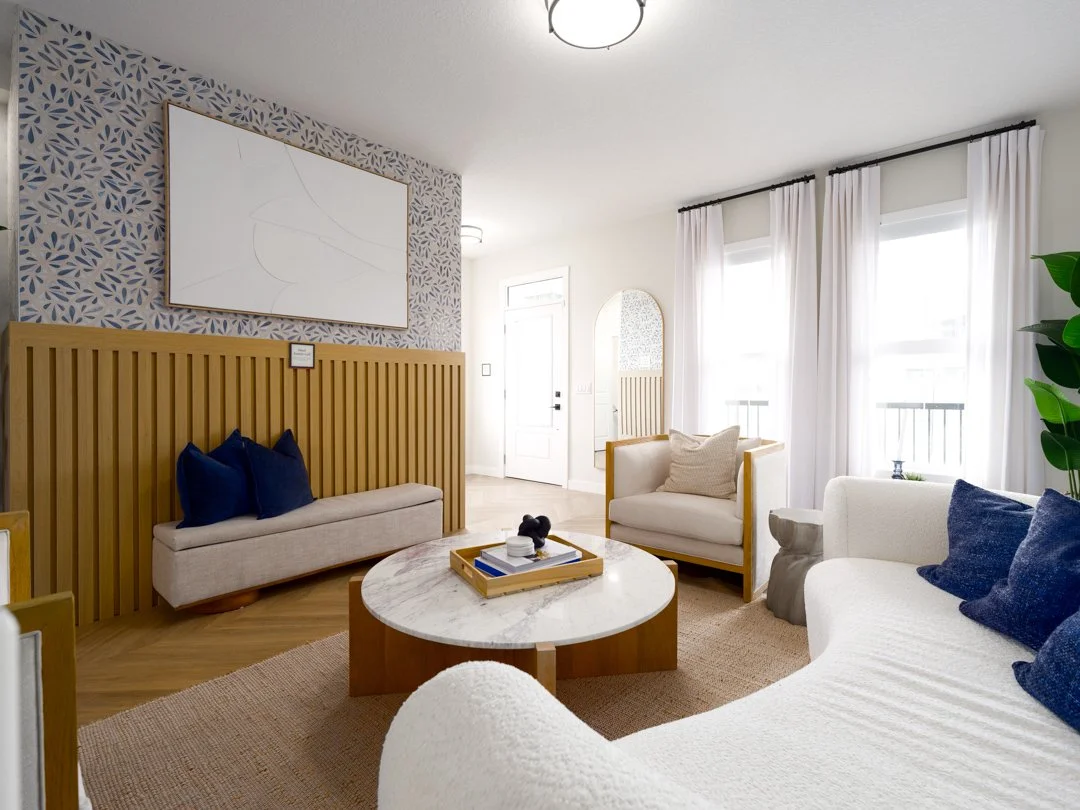 Living room with white sofas and armchairs, blue cushions, wooden coffee table with tray, large windows with white curtains, patterned wallpaper, and a minimalist art piece on the wall.