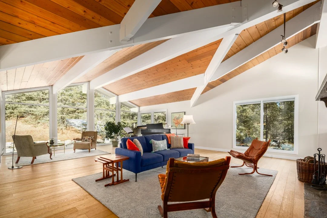 Living room with high vaulted ceiling and large windows showing outdoor trees, featuring hardwood floors, a blue sofa with colorful pillows, an orange armchair, a gray area rug, a piano, and various seating and decor.