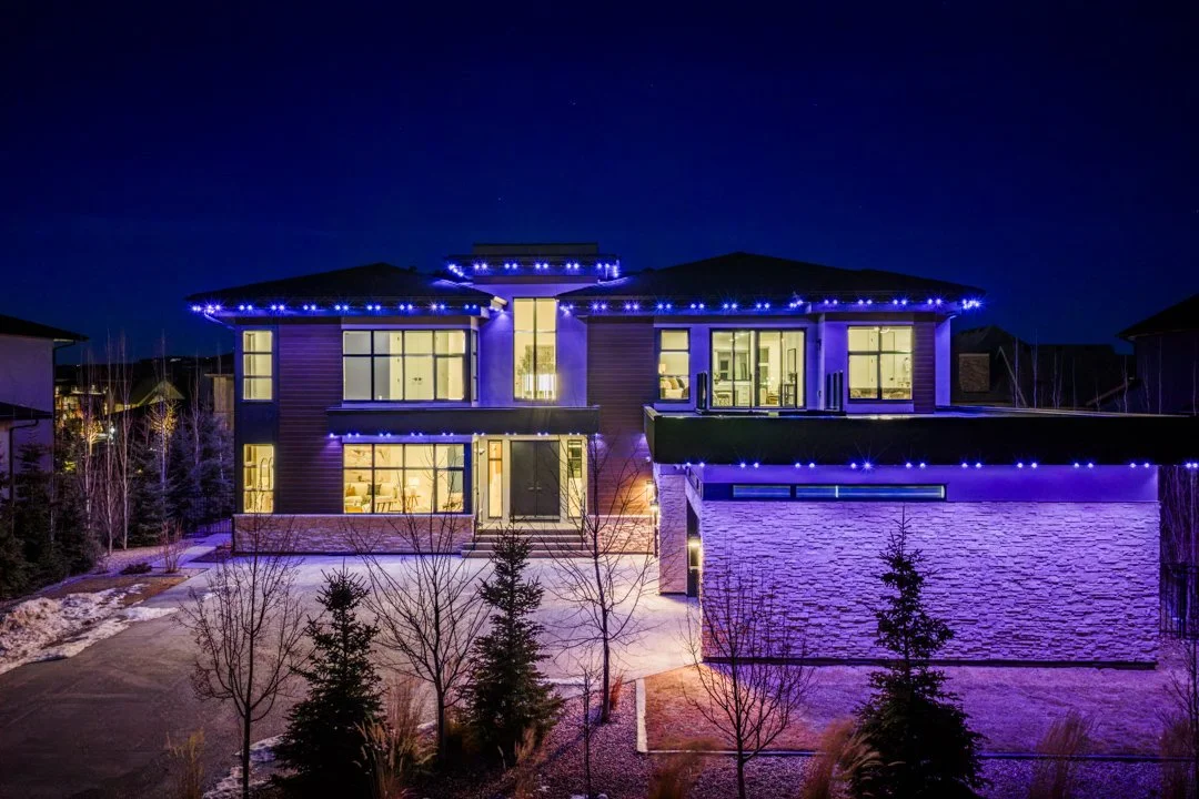 A modern, multi-story house illuminated at night with purple string lights outlining the roof, large windows, and a snow-dusted yard.