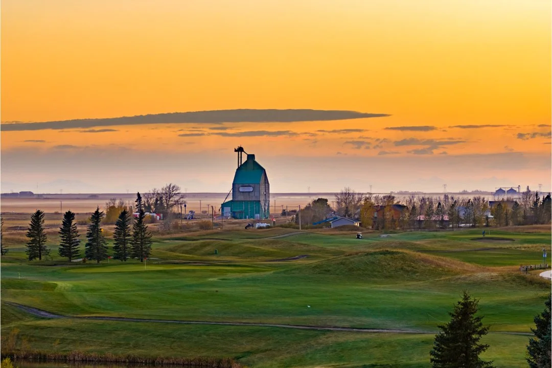 A scenic view of a golf course at sunset with a barn and some houses in the background.