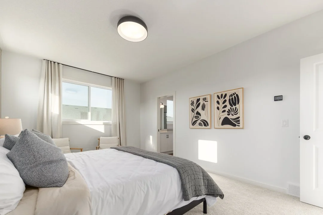 Bright bedroom with white walls, bed with gray pillows and blanket, window with white curtains, two chairs near the window, two abstract black and beige wall art pieces, ceiling light fixture.