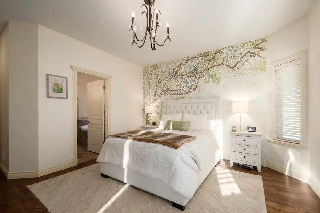Bedroom with a white bed, green and white pillows, wall mural of tree branches with blossoms, two white nightstands with lamps, window with blinds, hardwood floor, chandelier.