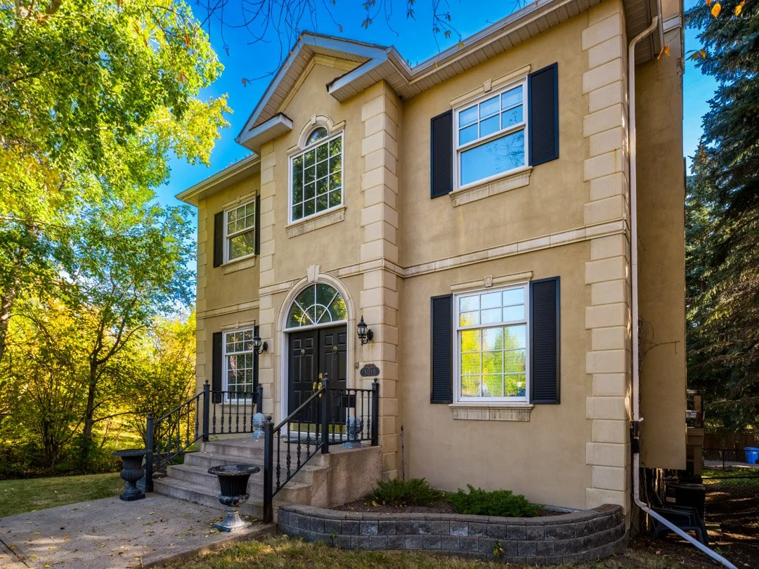 A three-story yellow stucco house with black shutters, black front door, arched window above door, and steps with black metal railings, surrounded by trees and a landscaped yard.