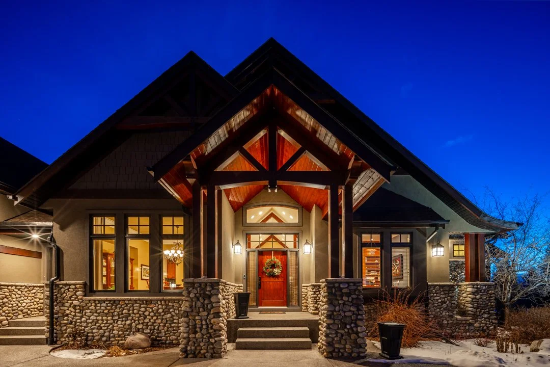 A front view of a modern house with a stone foundation, wooden accents, and large windows, illuminated at night with outdoor lighting, under a clear blue evening sky.