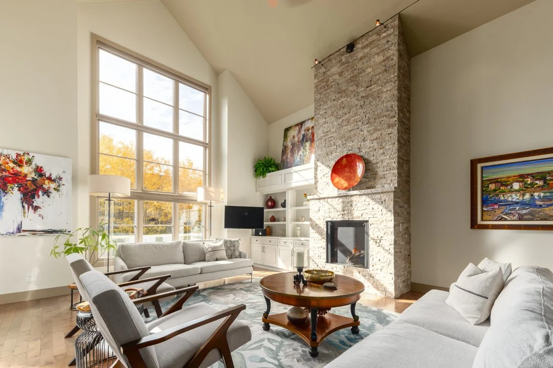 Living room with large window, white walls, beige sofas, wooden chairs, a coffee table, a stone fireplace with decorative art, hanging ceiling lights, art on the walls, and hardwood flooring.