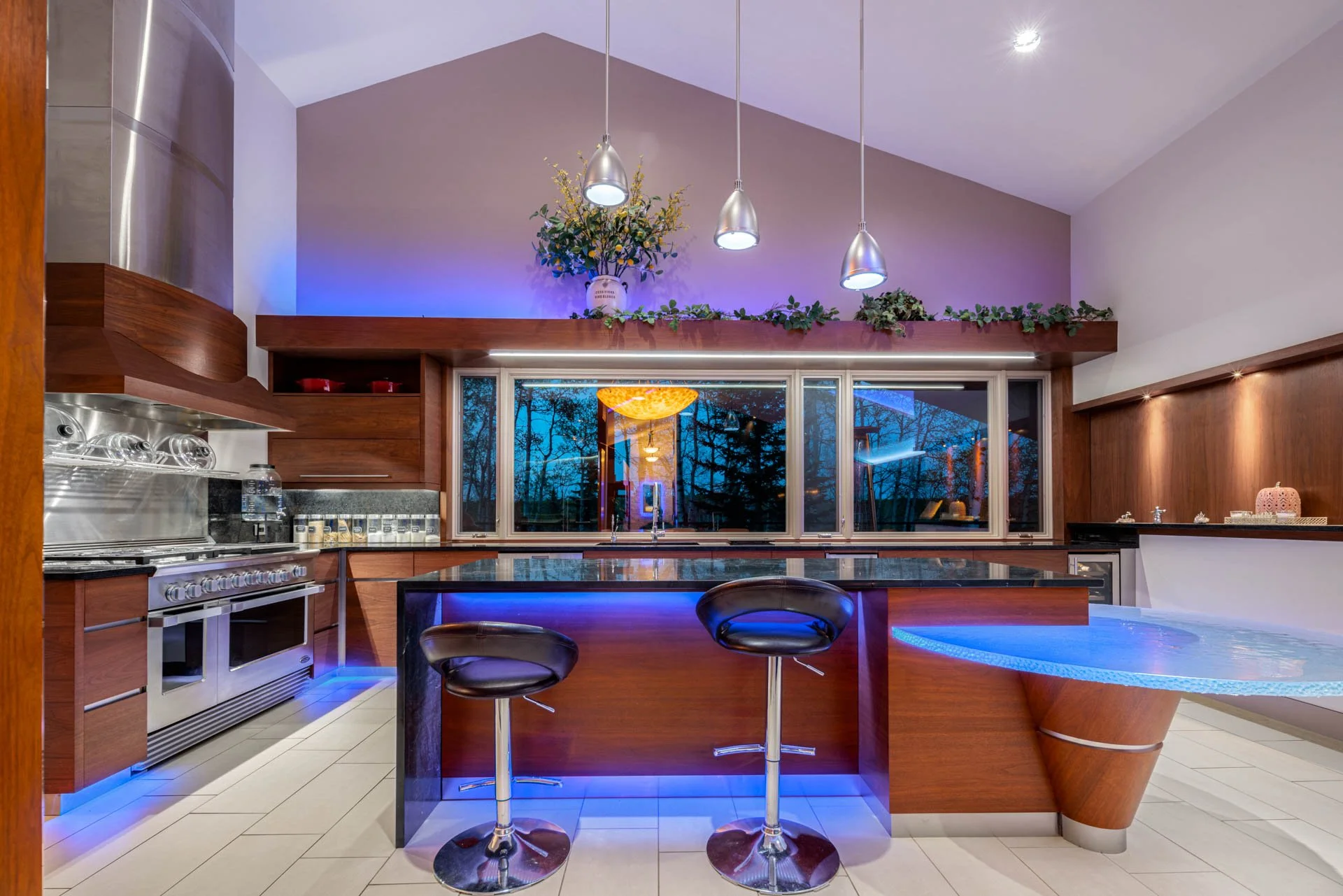 Modern kitchen with wooden cabinets, stainless steel appliances, a black kitchen island with blue accent lighting, and a large window showing trees outside. There are three pendant lights hanging from the ceiling, and decorative plants and flowers on