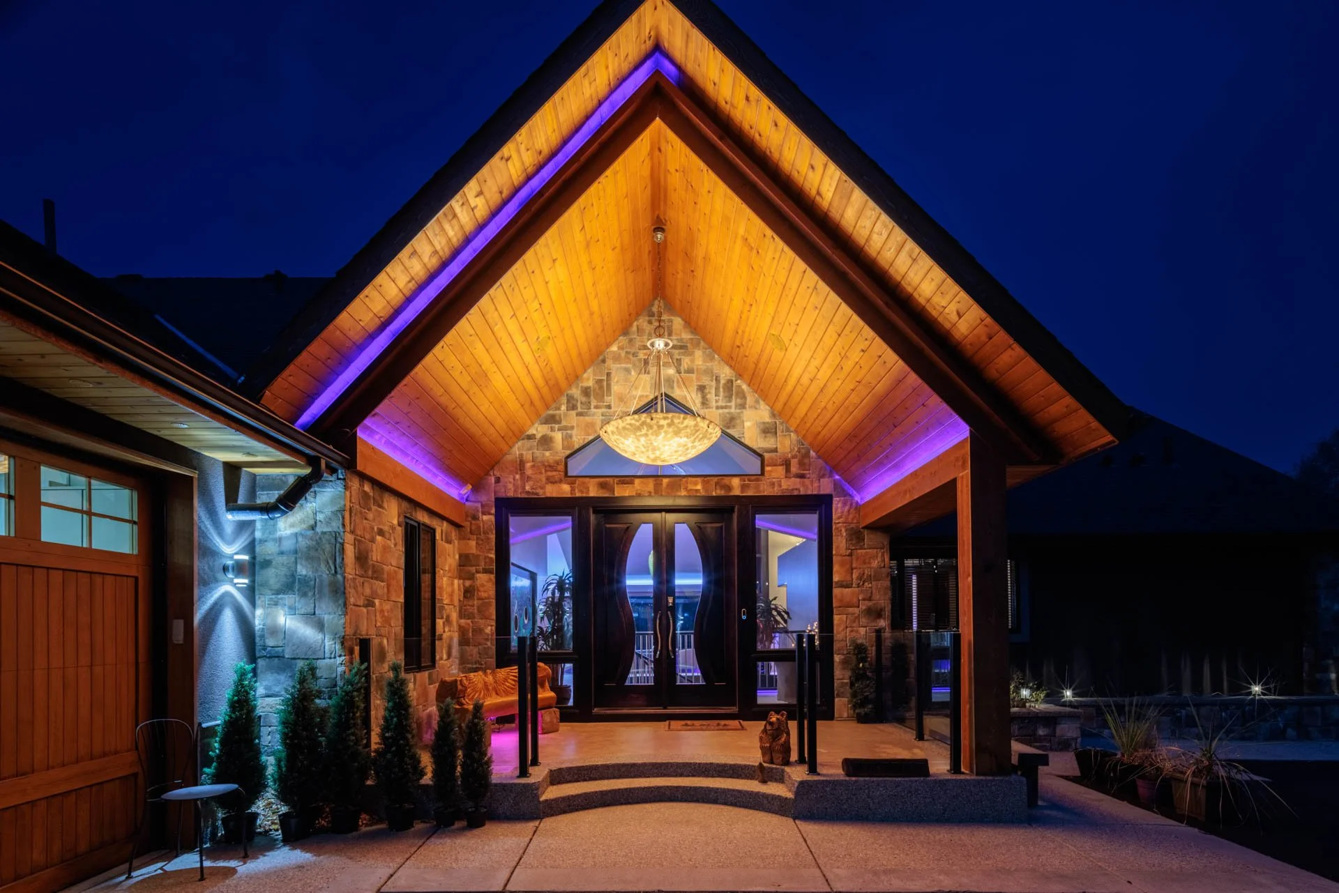 Night view of the front entrance of a house with a wooden A-frame roof, stone walls, illuminated by purple and yellow lighting, with a chandelier hanging inside, a small statue outside, and potted plants near the steps.