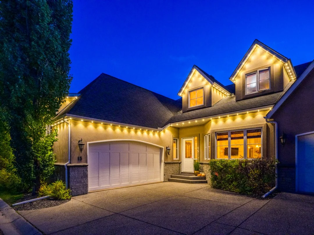 A house at night decorated with yellow string lights along the roofline, with a closed garage door, a front porch with steps, and windows with warm interior lighting.