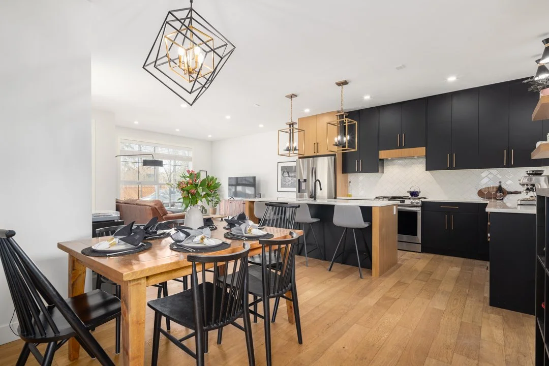 Modern kitchen and dining area with black and wood cabinetry, a wooden dining table set for six, pendant lights, and a living space in the background.