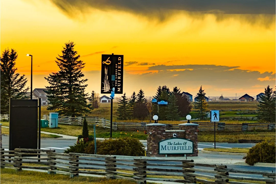 Sunset over a neighborhood with trees, houses, and signs for The Lakes of Muirfield development.