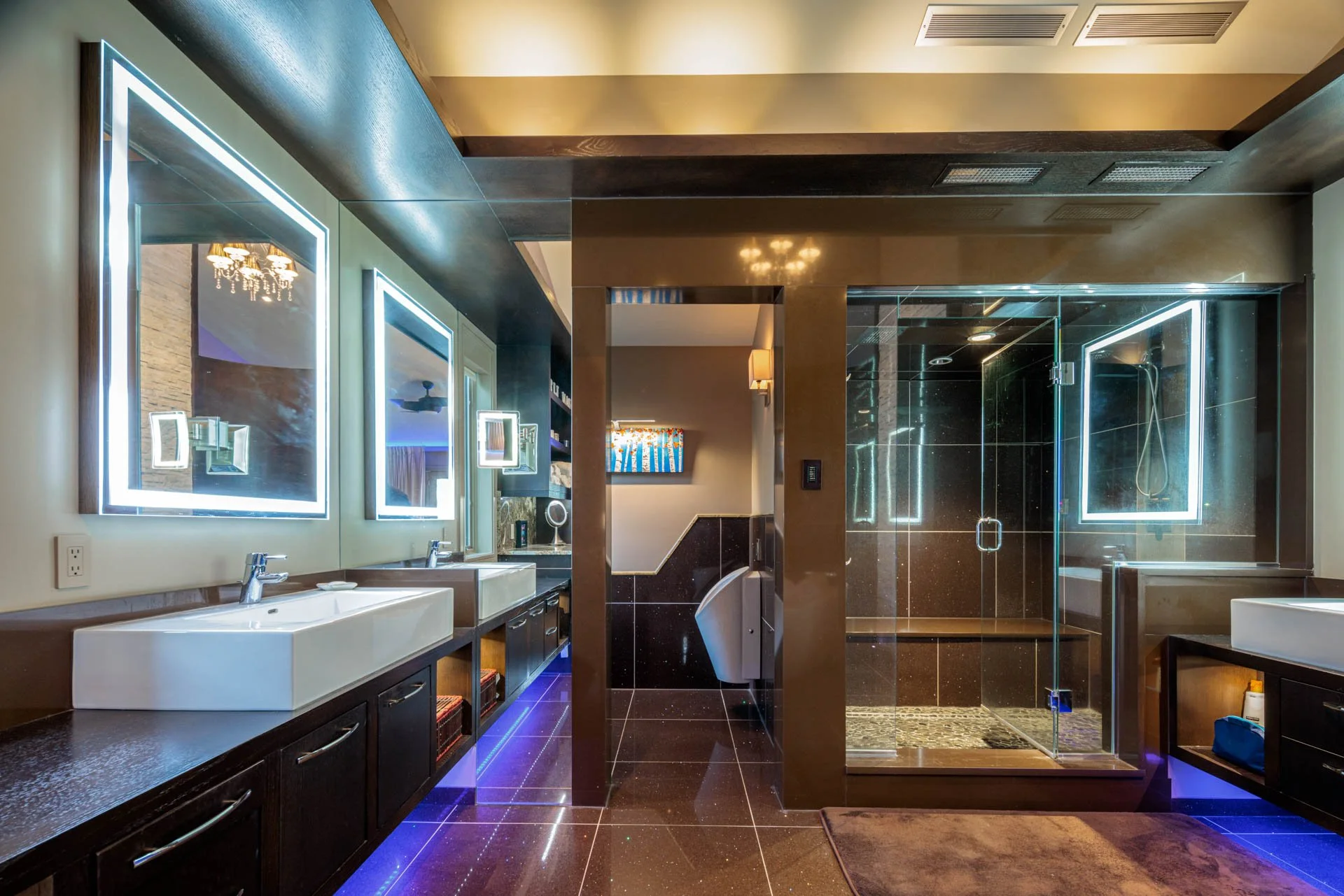 Modern bathroom with twin sinks, illuminated mirrors, a shower with glass walls, and dark tiles with blue LED lighting.