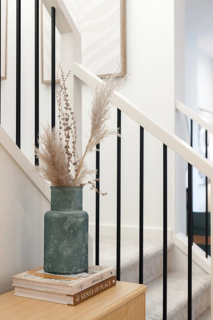 A large green vase with dried beige and brown pampas grass sits on a small wooden table, which has several books underneath, in front of a staircase with black metal railing.