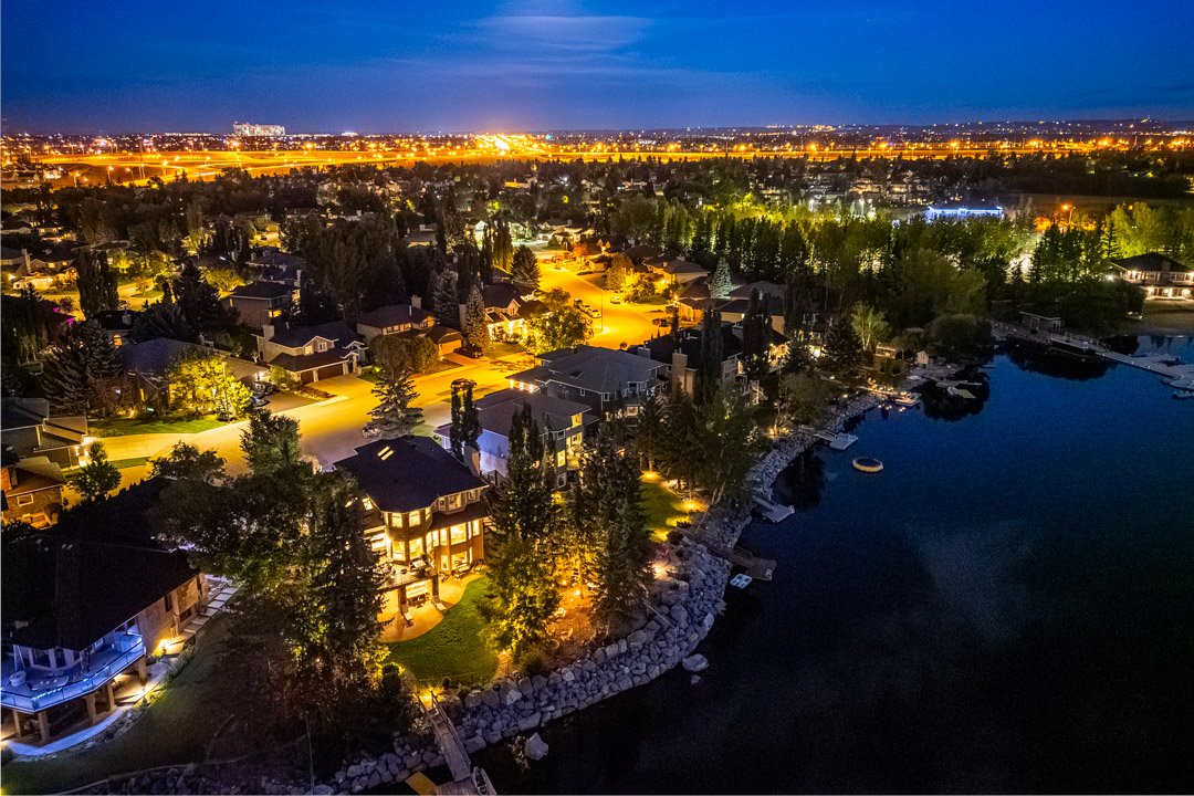 Nighttime aerial view of a lakeside neighborhood with illuminated houses and trees, a boat on the water, and city lights in the distance.