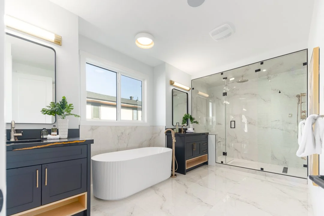 Modern bathroom with white marble floors and walls, a white freestanding bathtub, blue bathroom vanities with gold fixtures, a glass-enclosed shower with marble walls, and two windows providing natural light