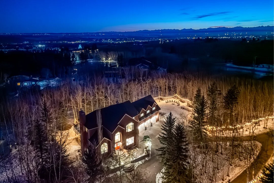 An illuminated house surrounded by trees in a snowy landscape at twilight with distant city lights in the background.