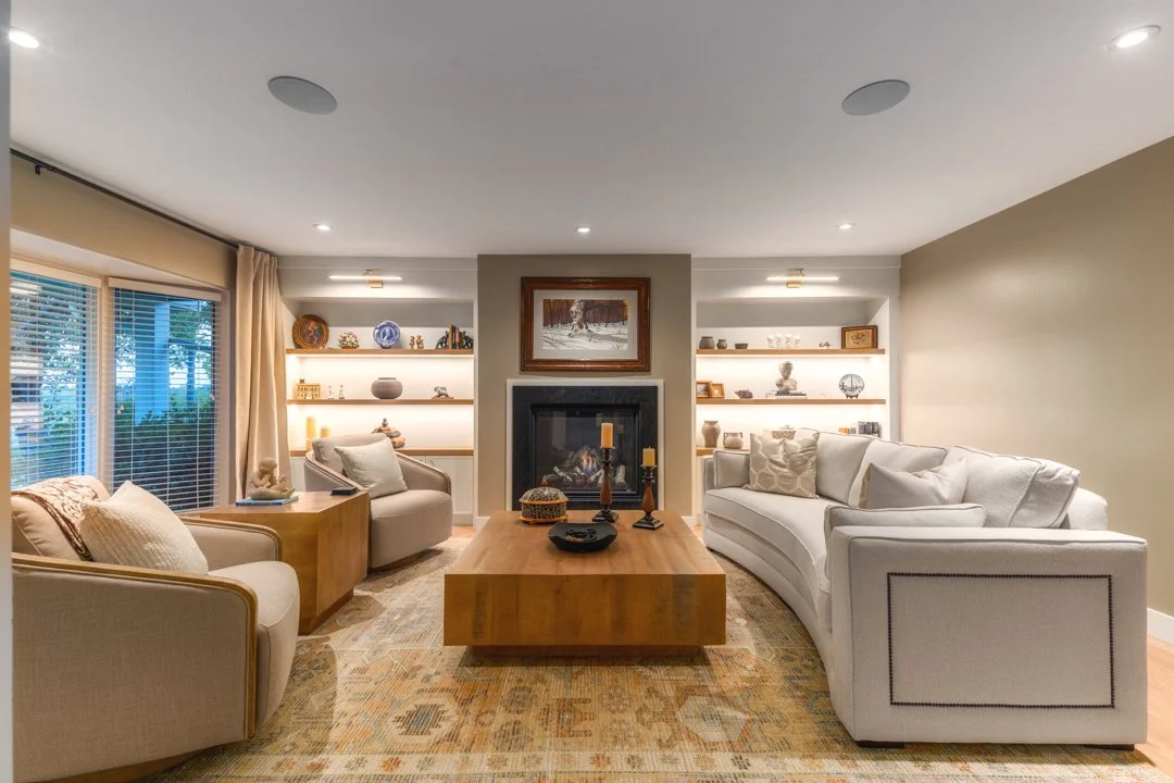 Living room with beige and white sofas, a wooden coffee table, built-in shelves with decorative items, a fireplace with a framed painting above it, large windows with blinds and curtains, and recessed ceiling lights.
