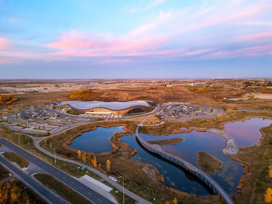 An aerial view of a modern building with a curvy roof, surrounded by parking lots, lakes, and roads in a rural landscape at sunset.