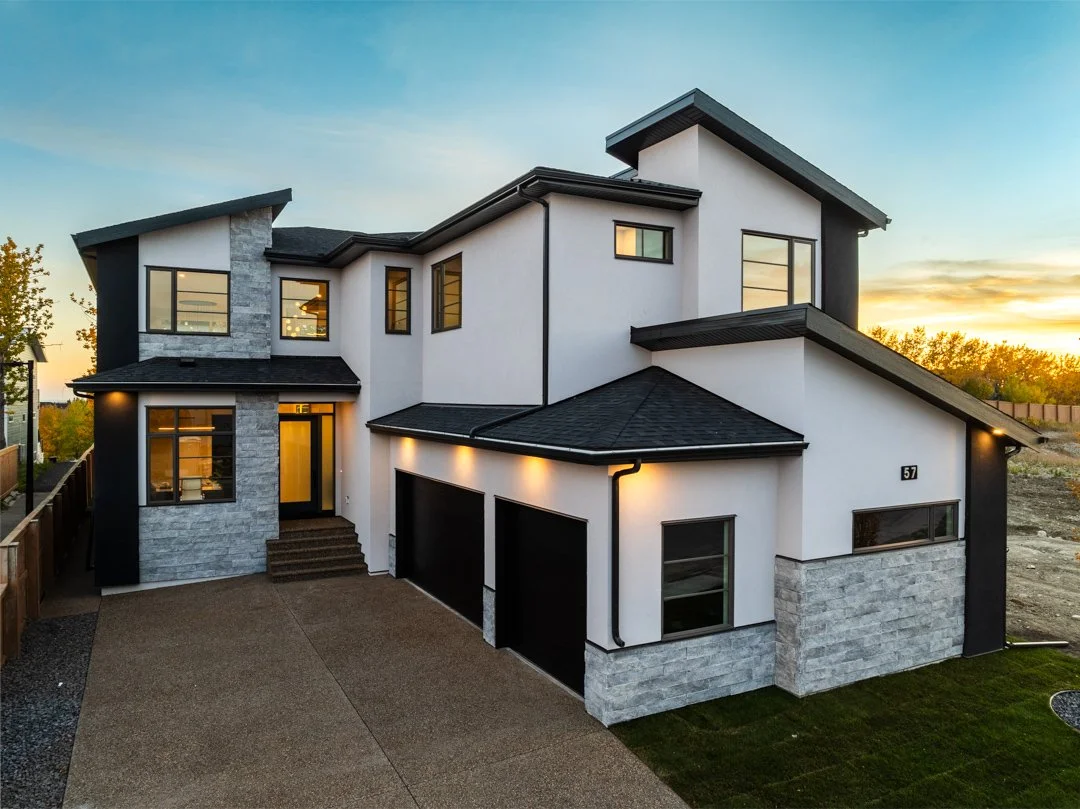 Modern two-story house with white walls, black trim, gray stone accents, multiple windows, and a three-car garage at sunset.