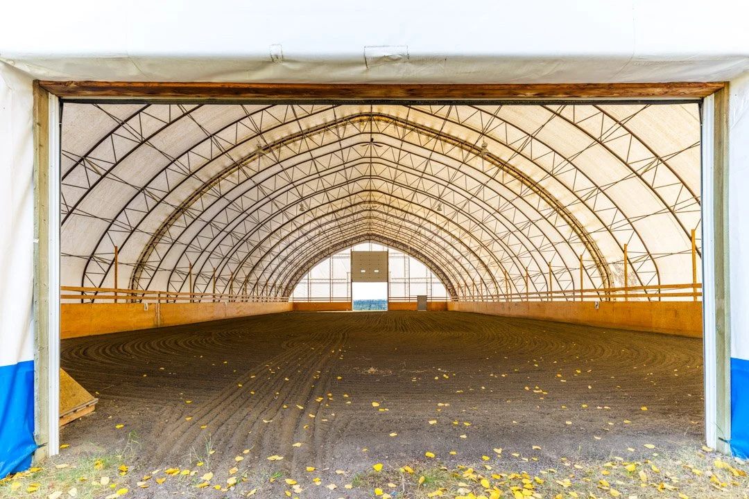 Indoor equestrian riding arena with a dirt floor, wooden barriers, and a fabric roof supported by metal framing, viewed from the entrance.
