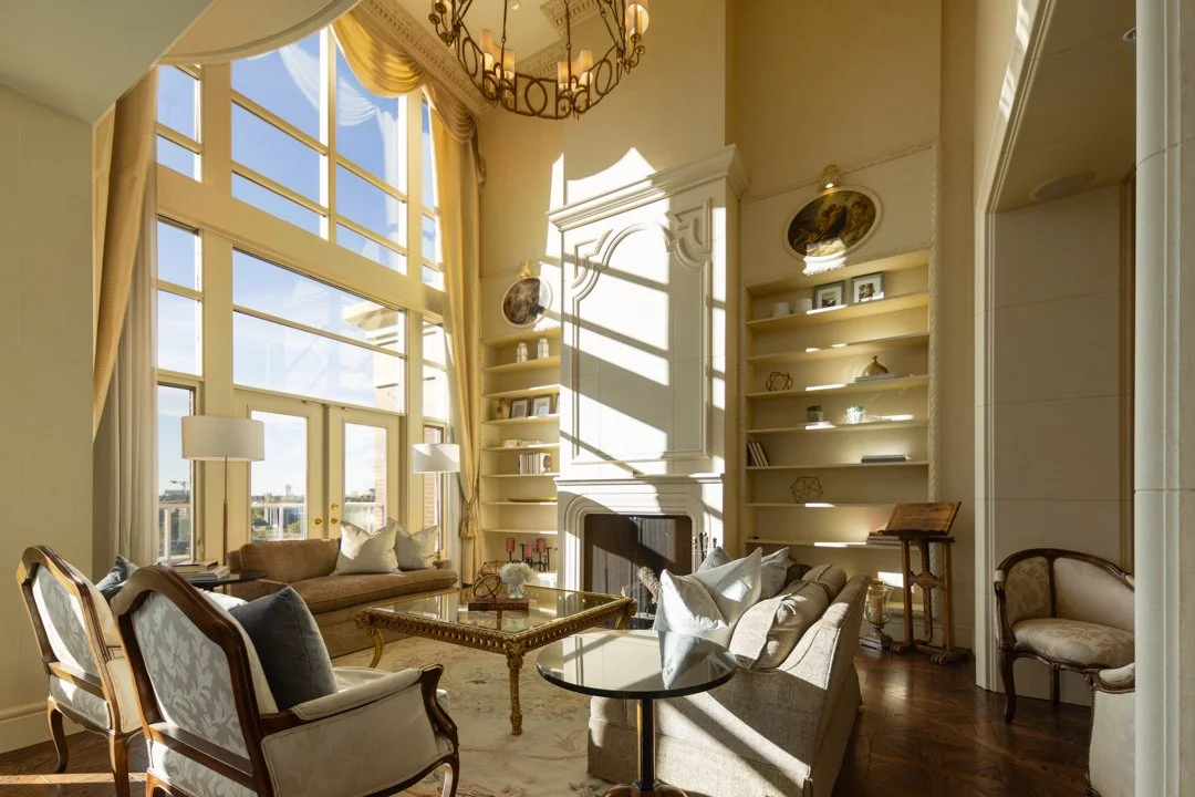 Elegant living room with tall windows, cream-colored furniture, a fireplace, built-in shelves, and a chandelier, illuminated by sunlight.