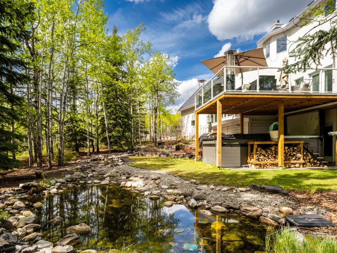 A house with a wooden deck, glass railing, and outdoor seating area, surrounded by lush green trees and a small pond with rocks.