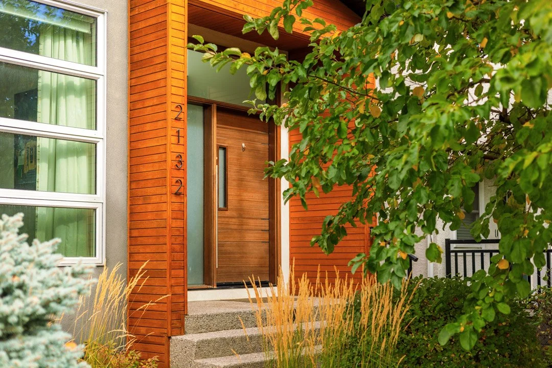 The image shows the front entrance of a modern house with a wooden door, a large window to the left, and a lush green tree partly covering the view. There are concrete steps leading up to the door, and decorative grasses and plants surround the entra