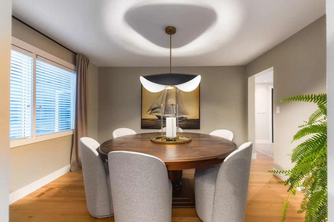 A dining room with a round wooden table, six gray upholstered chairs, a modern crescent-shaped ceiling light, a nautical-themed painting, a window with blinds, beige curtains, and a green potted plant.
