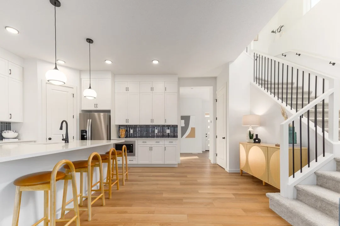 Modern kitchen with white cabinets, stainless steel appliances, a kitchen island with four wooden chairs, and a staircase with black railing and gray carpeted stairs.