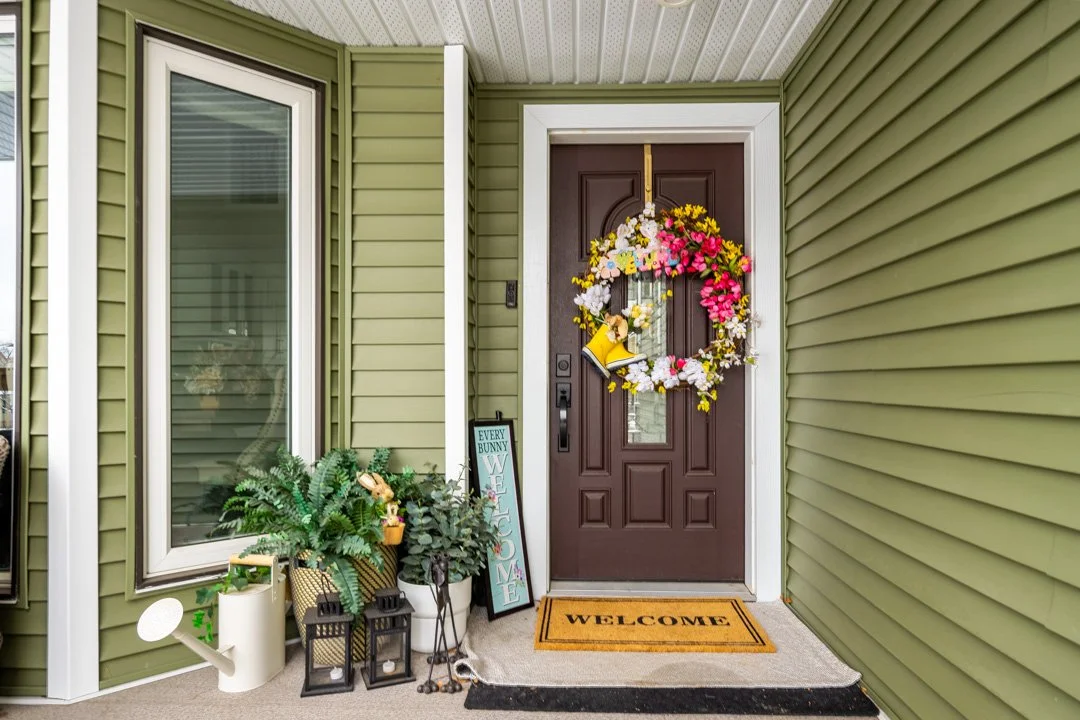 Front door decorated with a spring flower wreath, with potted plants, lanterns, and a welcome sign on the porch.
