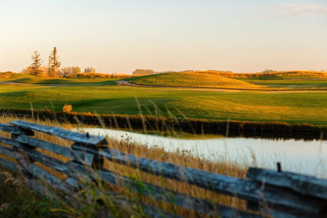 Scenic view of a golf course with rolling green hills, a water hazard, and trees in the distance, under a clear sky.