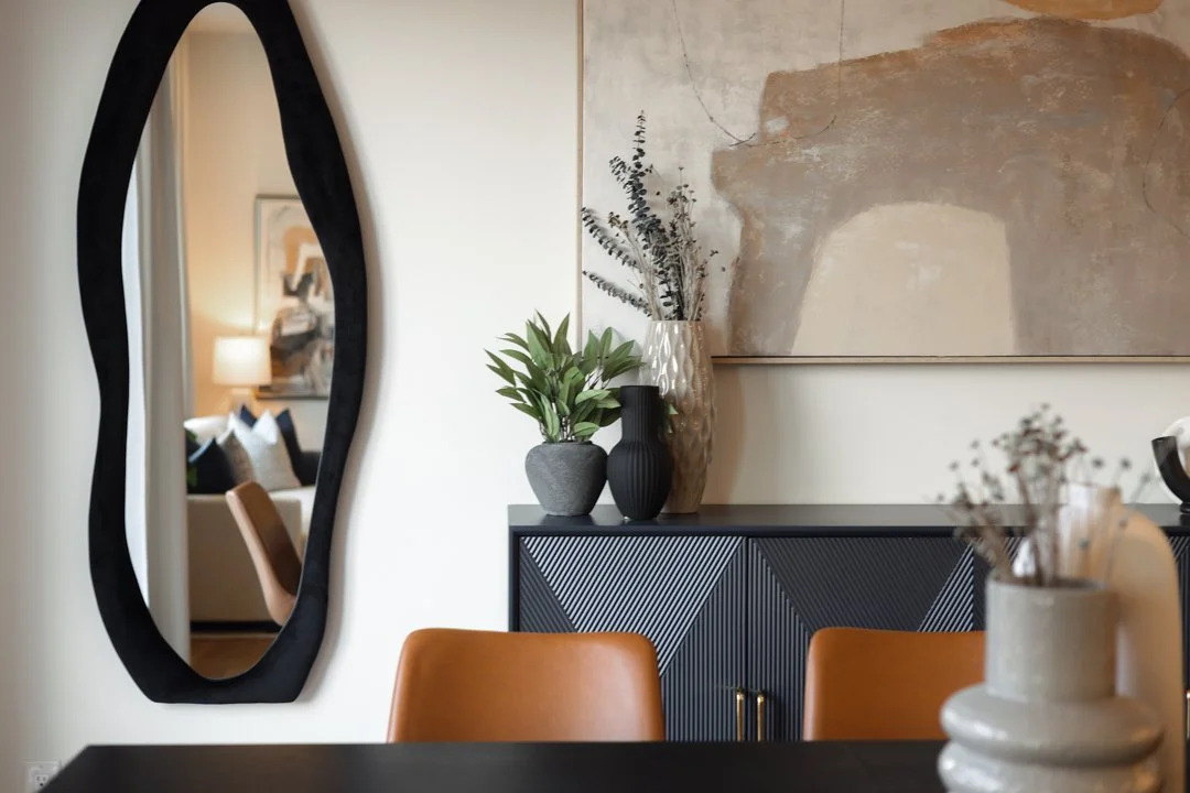 A modern dining room with a black oval mirror, a black sideboard with angled, textured doors, various vases with plants on top, a large abstract wall art, and tan chairs around a dark dining table.