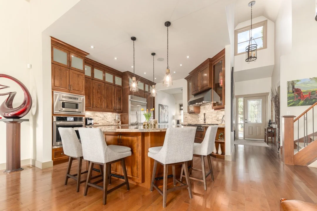 Modern kitchen with wooden cabinets, granite countertops, and a central island with four upholstered chairs, pendant lights, and stainless steel appliances.