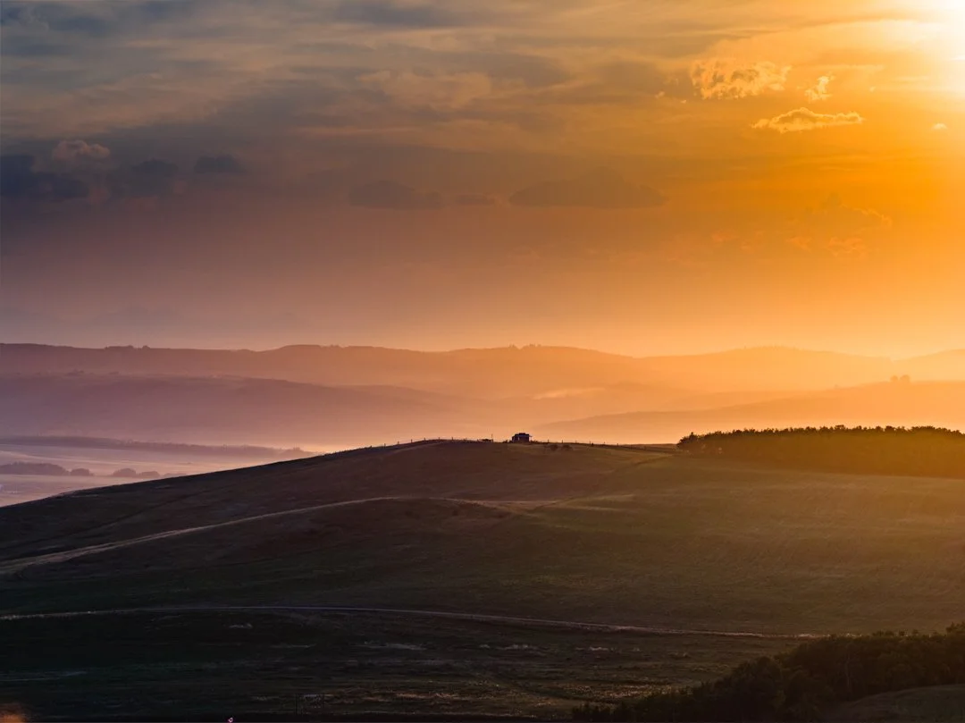 Sunset over rolling hills with layered mountains in the distance and a small building on the hilltop.