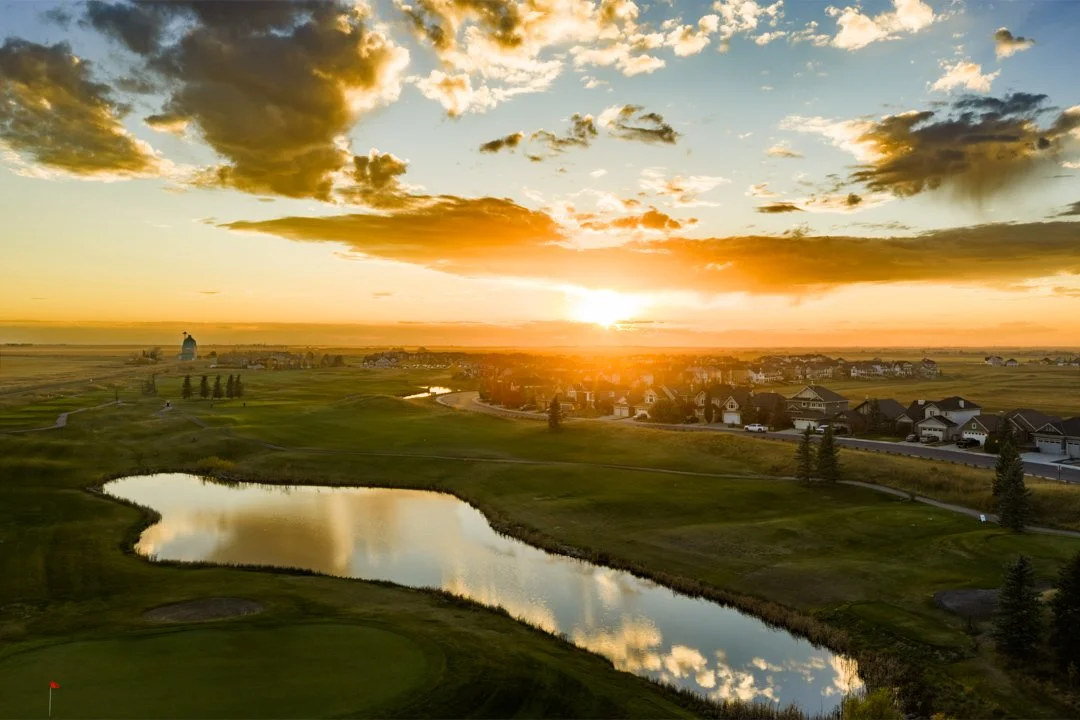 Sunset over a suburban landscape with a golf course, small water bodies, houses, and a cloudy sky.