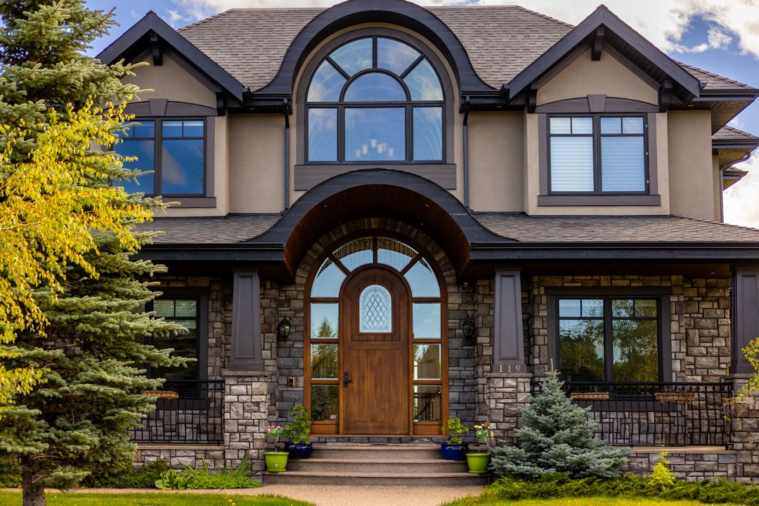 Front view of a two-story house with a stone exterior, arched wooden front door, large second-story window, and landscaped yard with trees and potted plants.