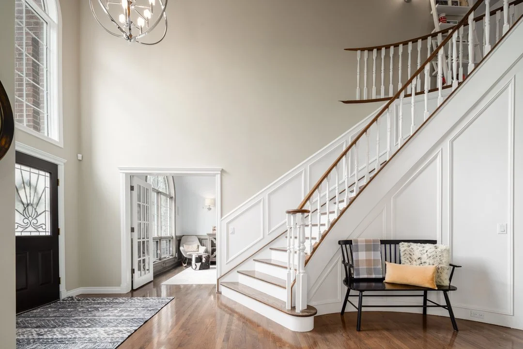 Interior of a house with a curved staircase, black bench with pillows, hardwood floors, and large windows.