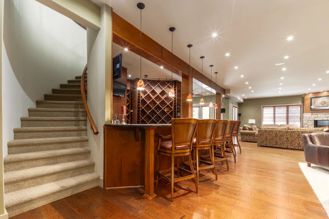 Living room with a staircase, bar area with four barstools, and a seating area with sofas and a stone fireplace.