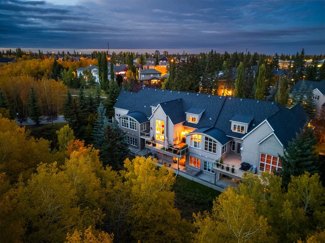 An aerial view of a large, multi-story house with illuminated windows at dusk, surrounded by colorful autumn trees and neighboring houses in a suburban neighborhood.