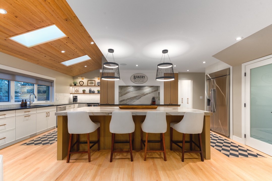 Modern kitchen with a central island, four white chairs, wooden countertops, and a skylight ceiling, with a bakery sign on the wall.