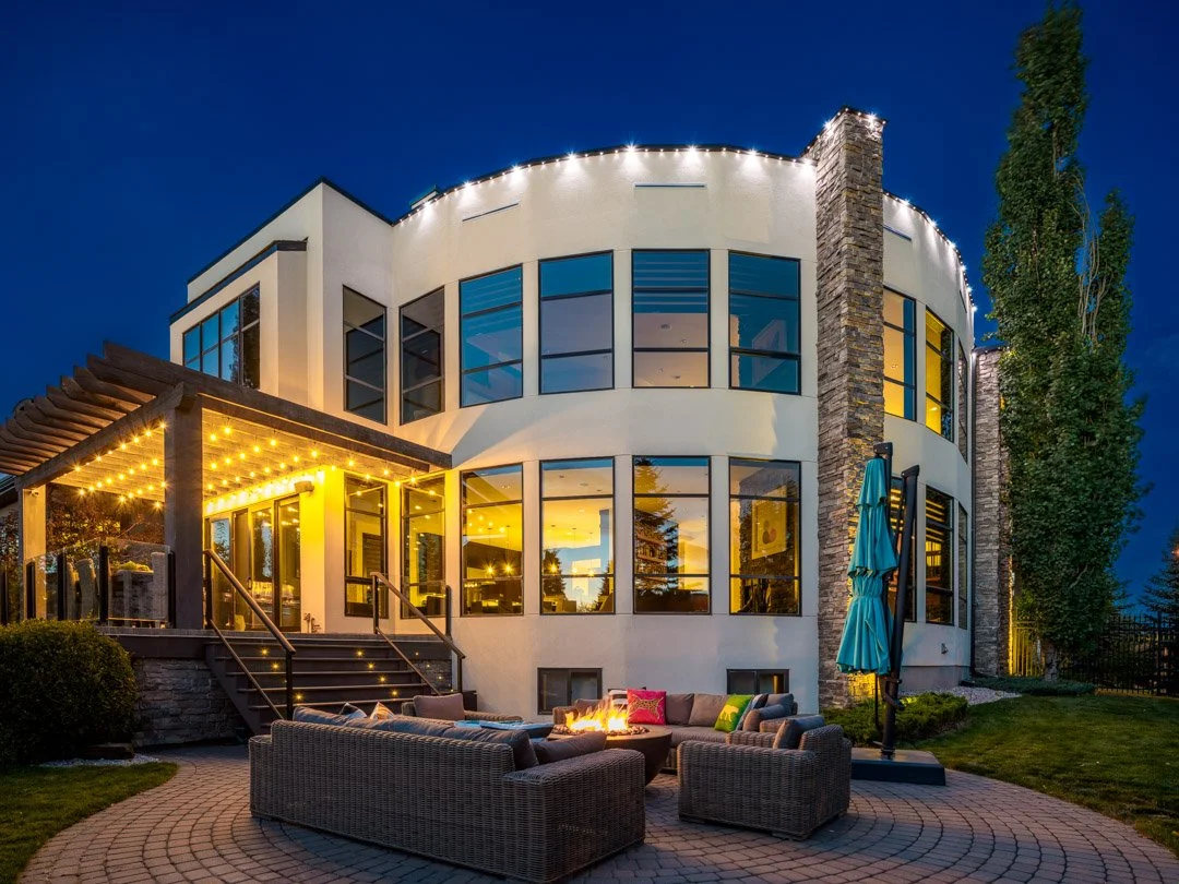 Modern two-story house with large windows, outdoor seating area, fire pit, and string lights at dusk.