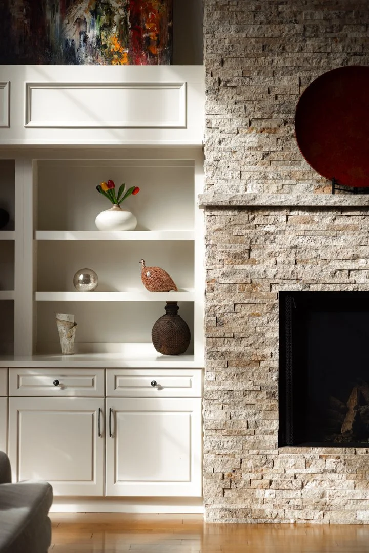 A living room with a white built-in shelf displaying decorative items, including a white vase with tulips, a ceramic bird sculpture, a glass orb, and vases, beside a stone fireplace with a red decorative plate above it. There is an abstract colorful 