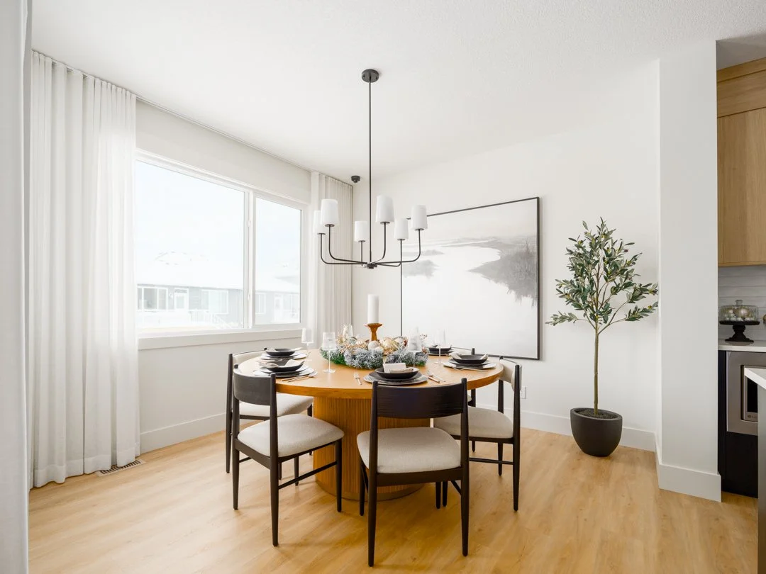 A modern dining room with a round wooden table set with black plates, wine glasses, and holiday decorations, surrounded by four black and beige chairs. There's a large window with white curtains, a black chandelier hanging above the table, a large ab