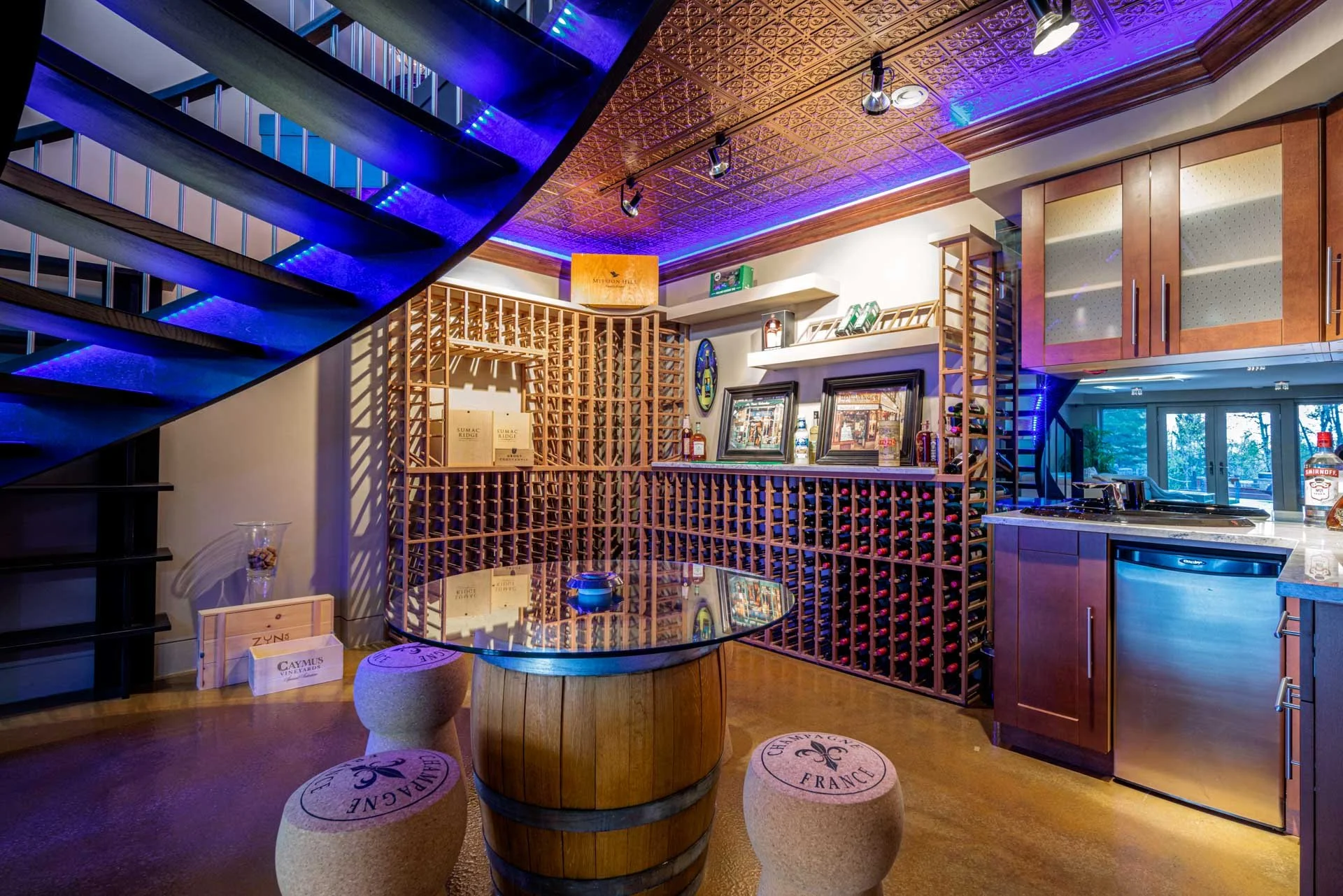 A cozy wine cellar or tasting room with a wooden wine rack, a glass-topped table, cork stools, and decorative bottles and framed pictures on the shelves. There's a staircase with blue lighting and a kitchen counter with a sink and window in the backg