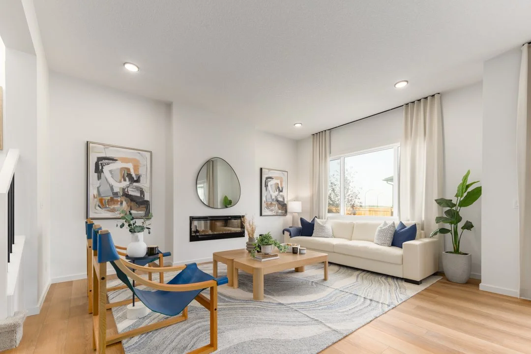 Bright, modern living room with white walls, large window with white curtains, white sofa with blue and white pillows, abstract artwork on walls, round mirror, fireplace, coffee table with books and plants, wooden chairs with blue cushions, hardwood 