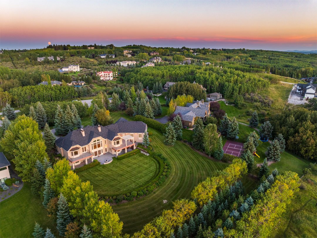 Aerial view of a large house with a well-maintained lawn and numerous trees, surrounded by other houses and lush greenery, during sunset.