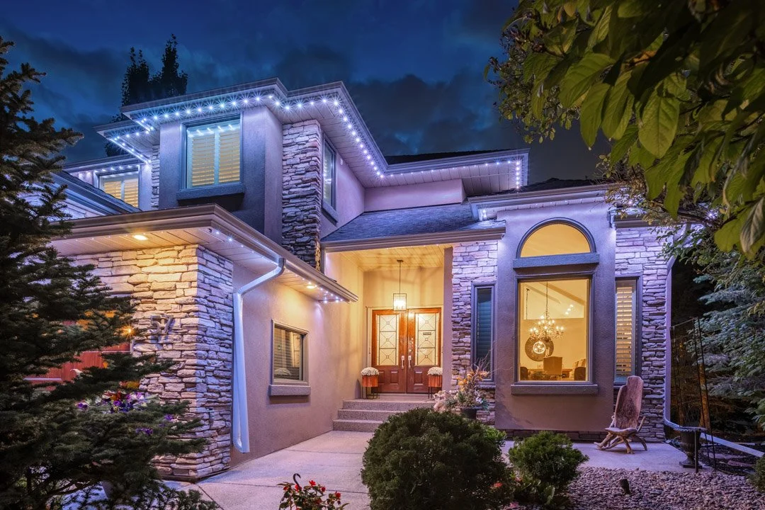 A modern two-story house with stone and stucco exterior, illuminated by exterior and interior lights at dusk, with a front porch, steps, plants, and trees surrounding it.