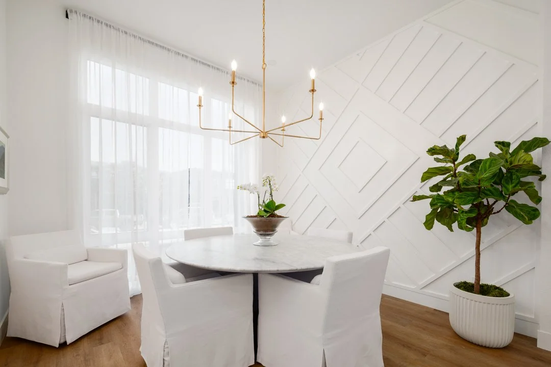 A bright, modern dining room with white walls, sheer curtains, a white round table, four white chairs, a gold chandelier, a potted plant, and a white textured accent wall.